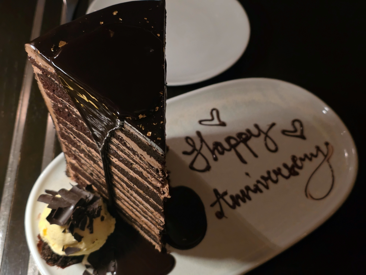 A slice of chocolate cake on a plate with "Happy Anniversary" written in chocolate syrup