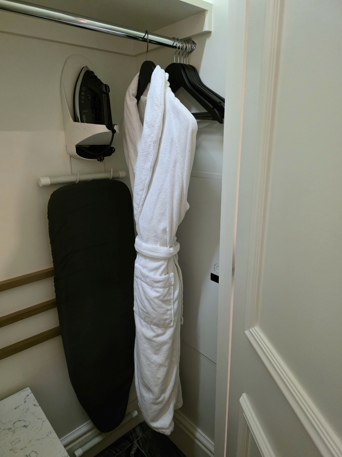 Open hotel closet hanging plush white bathrobes and an ironing board
