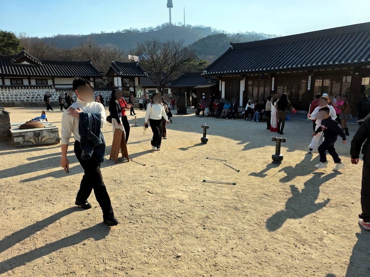 A view of the main square at Namsangol Hanok Village, where visitors are playing the traditional Korean game of Tuho (arrow-throwing). In the foreground, a person is about to throw a stick into a large ceremonial jar on a stand. Other people are watching, and traditional houses line the background.